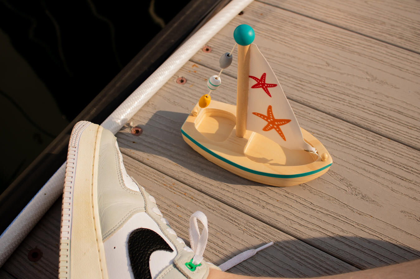 Wooden toy sailboat with starfish sails, colorful bead garland, and partially visible Air Force 1 sneaker on a wooden dock with dark water in the background.