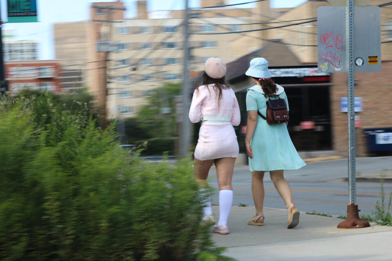 Two young women, dressed in fashionable pastel outfits, walk along a city sidewalk. The woman on the left wears a pink plaid skirt suit and a beret, while the woman on the right wears a light blue dress and a bucket hat, and carries a small brown backpack.