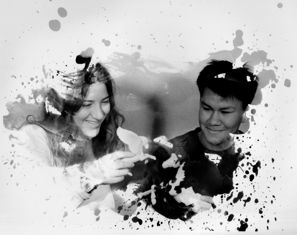 A black and white photo of two people smiling and playing with paper birds. Ink splatters overlay the scene, adding a creative, joyful feel.
