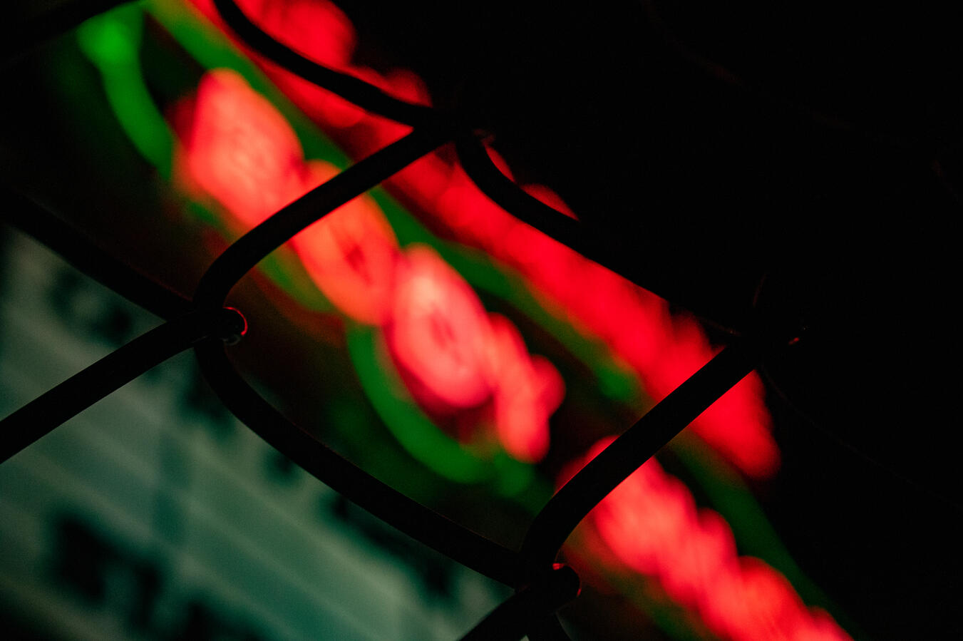 Abstract blurry red neon sign with green accents behind a dark wire fence.