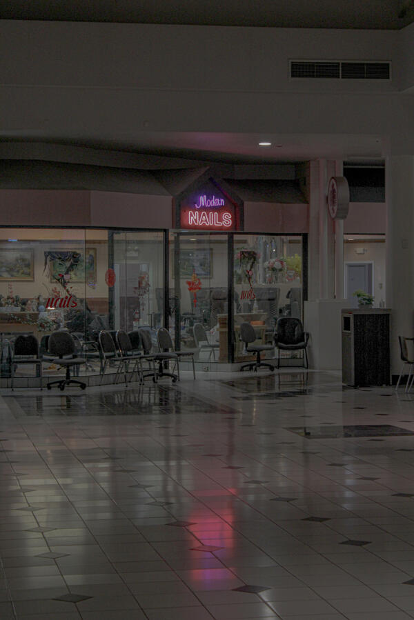 Neon sign reading "Modern Nails" above the entrance to a nail salon inside a dimly lit shopping mall, with rows of empty chairs in the waiting area.