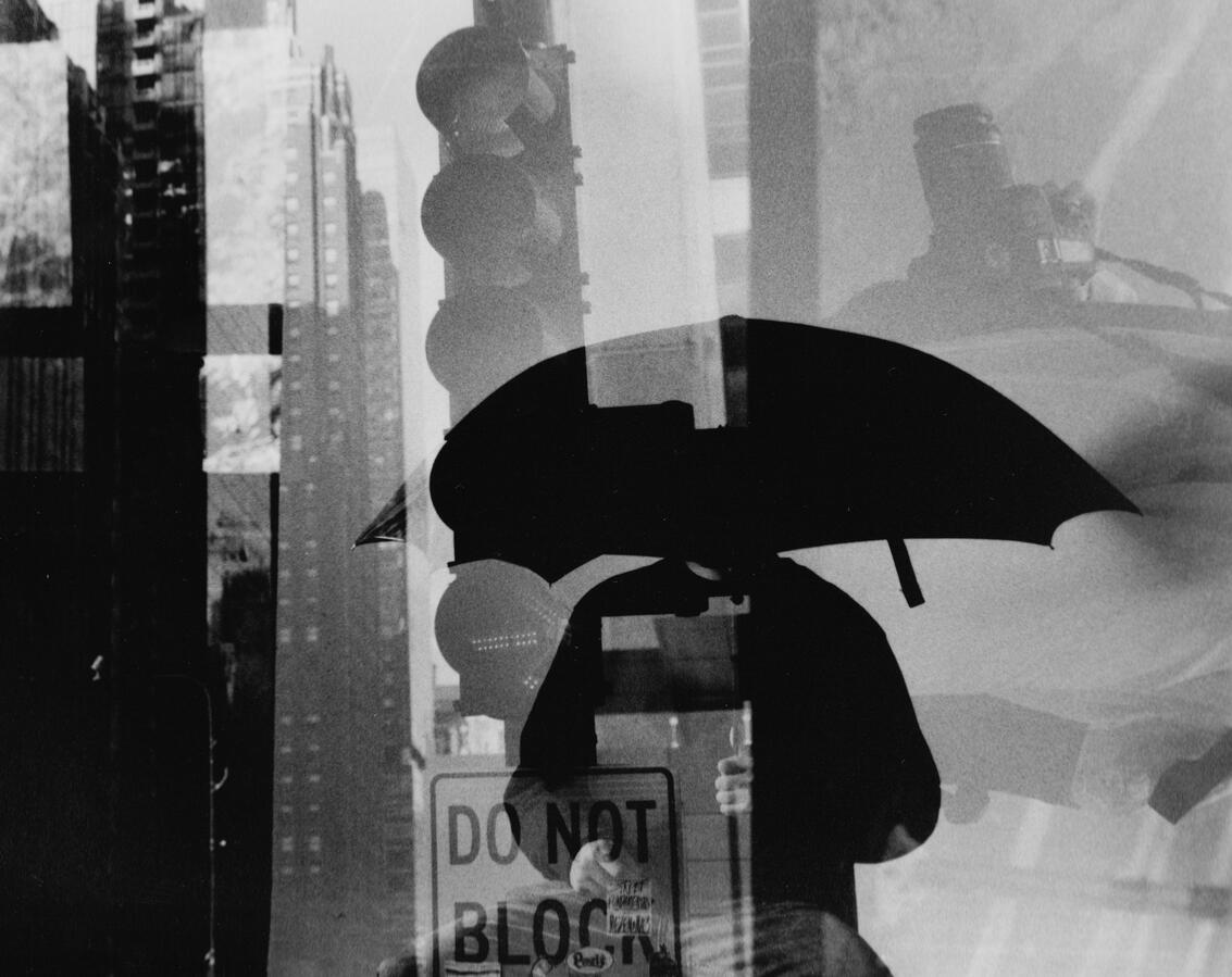 Abstract double exposure of a person holding an umbrella on a city street with a traffic light and buildings.