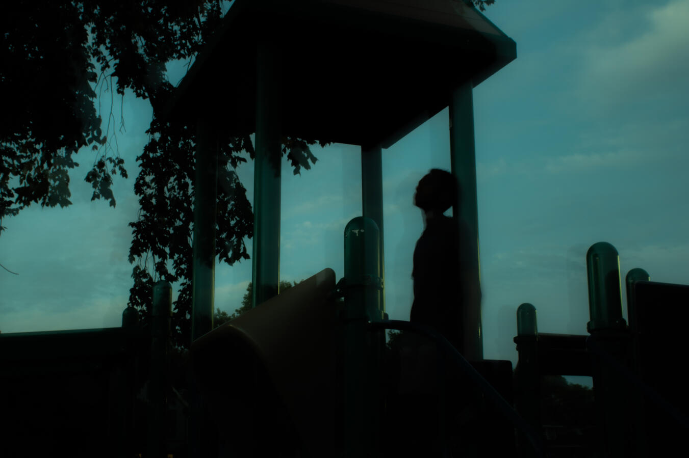 Silhouette of a person leaning against a pillar on a playground structure against a cloudy sky.