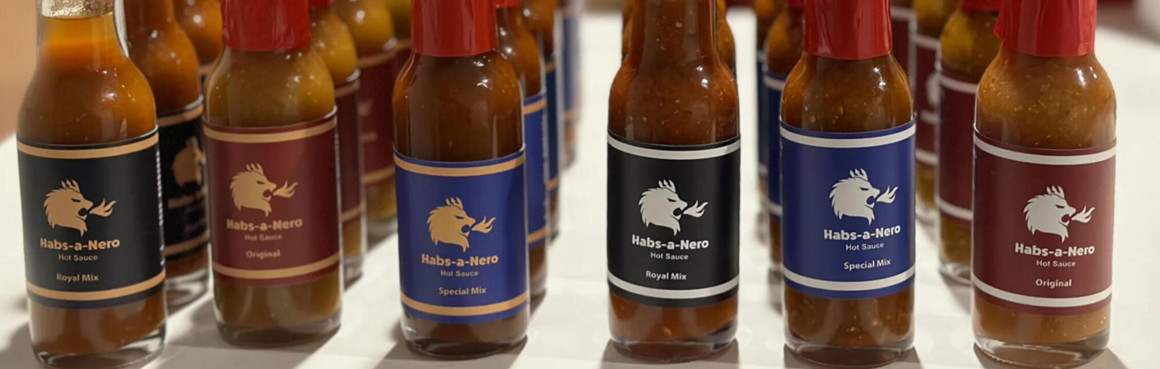 Rows of clear glass hot sauce bottles, labeled "Habs-a-Nero Hot Sauce". Some bottles have black labels, others have maroon or blue, and are sealed with red paper. The labels feature a gold illustration of a lion head breathing fire.